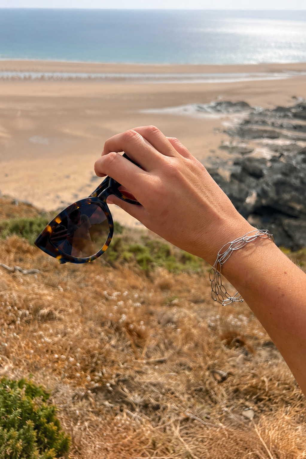 Henriette portée en bracelet, lunettes de soleil à la main devant la plage, JACQUAND