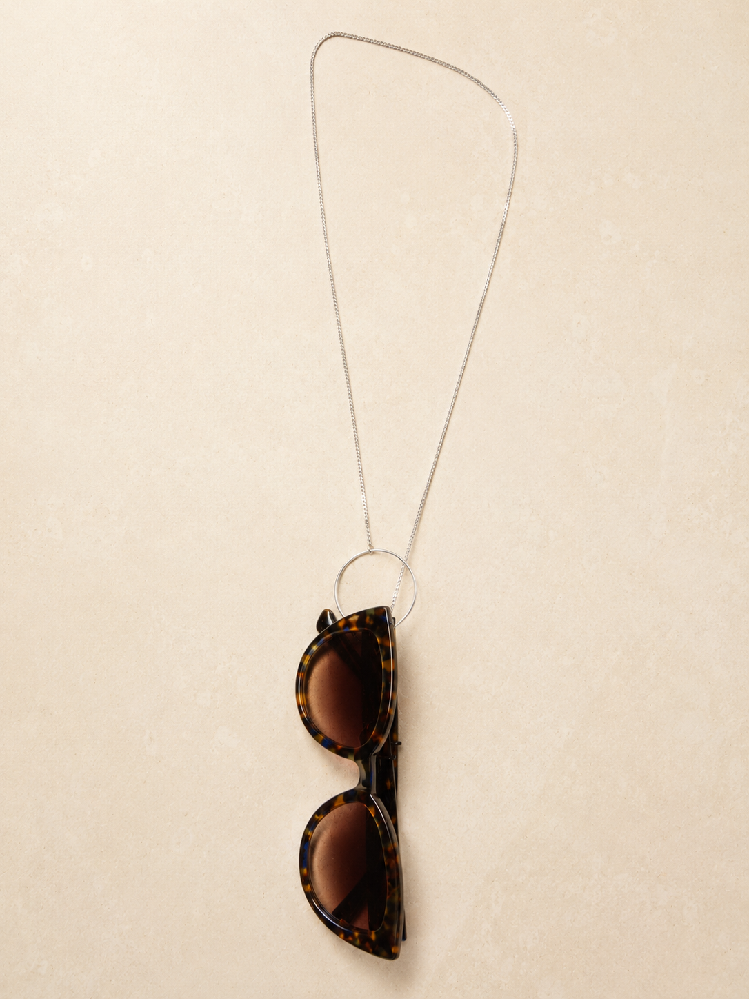 Azélie, chaîne argentée dont l'anneau retient une paire de lunettes de soleil écaille, posée à plat, JACQUAND