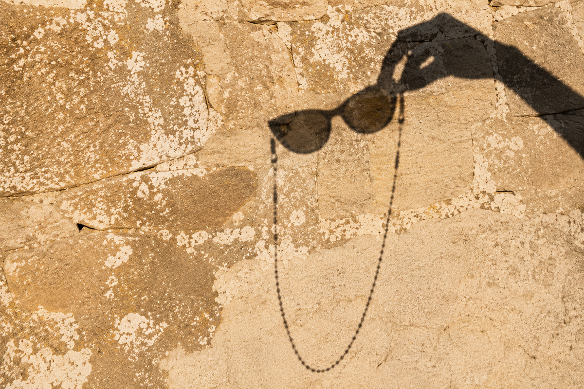 Ombre d'une main tenant des lunettes de soleil et leur chaîne JACQUAND, projetée sur un rocher au soleil